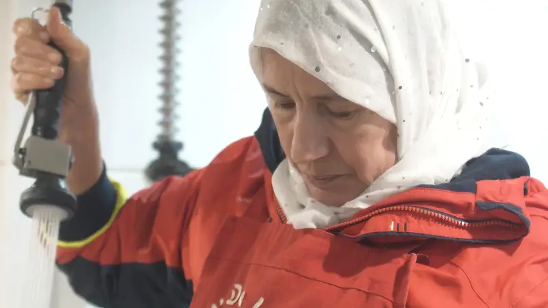 Une femme au foulard blanc et veste rouge, sous une douchette de cuisine.