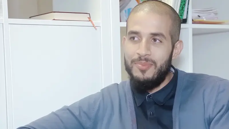 Un homme barbu, assis dans un bureau aux étagères blanches, pendant un entretien.